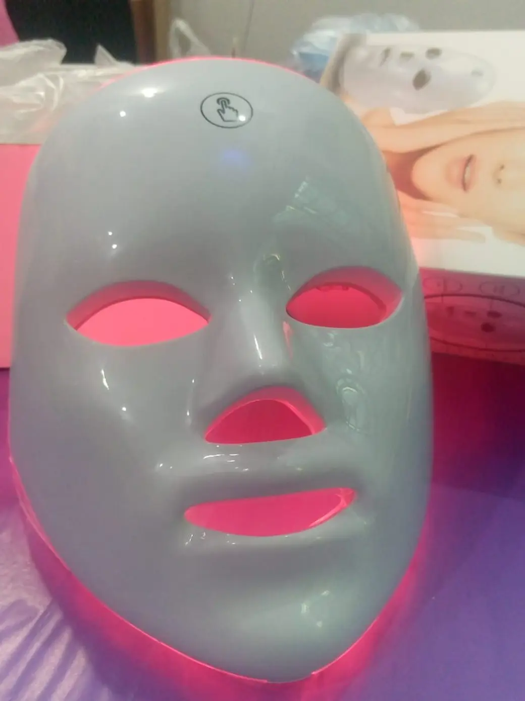 照片，一張亮著粉紅色燈的白色 LED 美容面罩放在桌上，後方是產品包裝盒。