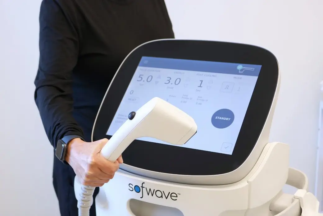 器材照片，一位穿深色衣服的人握著 Sofwave 的治療手把，後方是儀器觸控螢幕，上面顯示 5.0、3.0、1 等參數與 STANDBY。