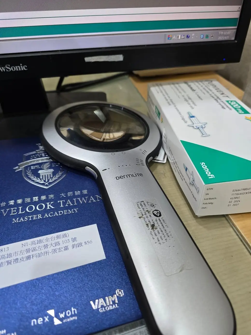 桌面照片，一支 DermLite 皮膚鏡放在深藍色的學院資料夾上，旁邊是一盒賽諾菲的預充填針劑藥盒，背景是電腦螢幕。