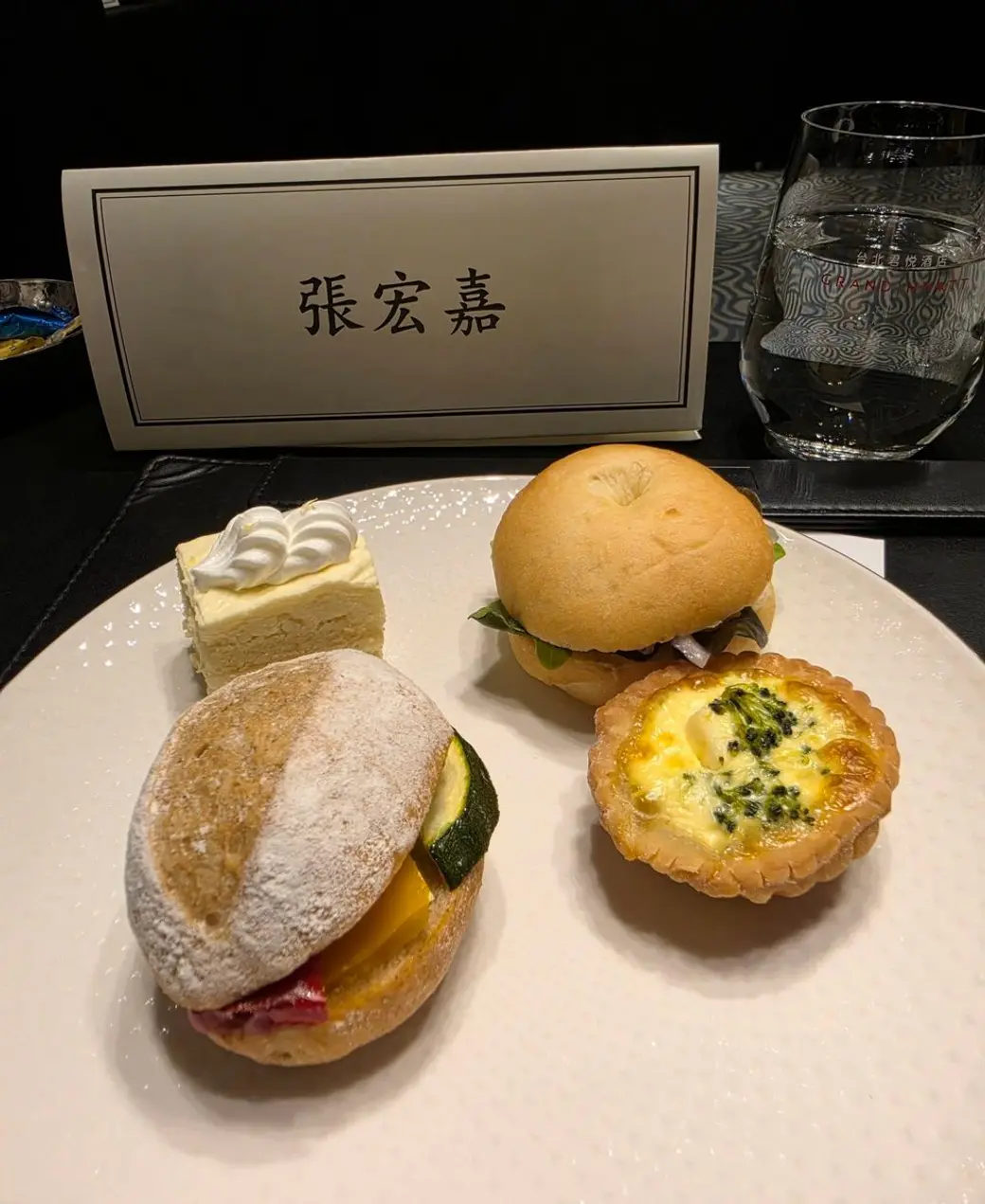 餐桌照片，桌上放著一張印著中文姓名的席卡，前方盤子裡是兩個麵包、一塊蛋糕與一個鹹派。