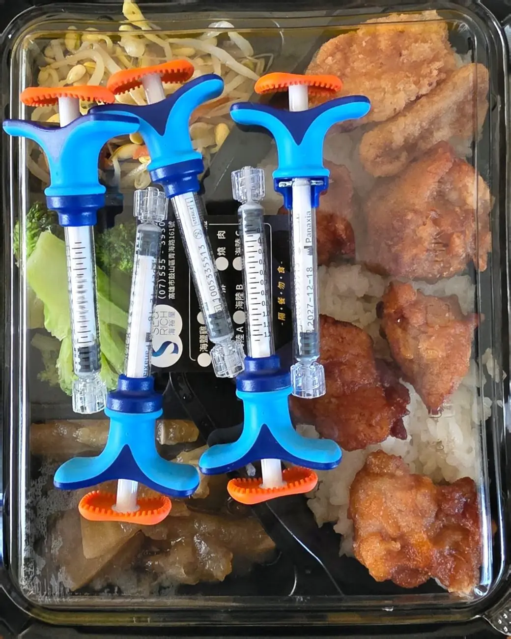 照片，一個便當盒上放著五支預充填的注射針筒，盒內是白飯、炸雞與配菜。