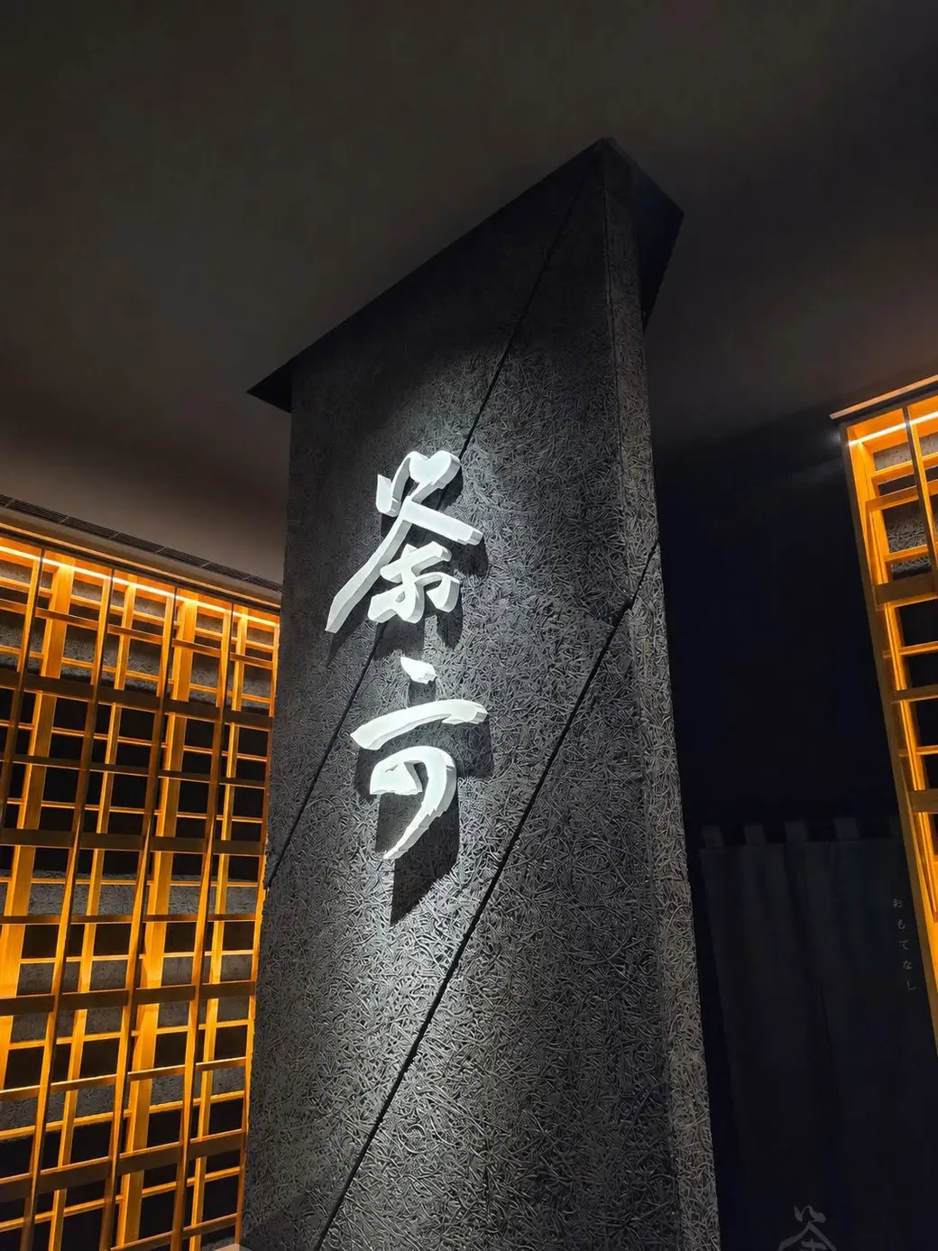 夜裡的店門口，深色石柱上有發光的店名招牌，兩側是暖黃的木格燈。