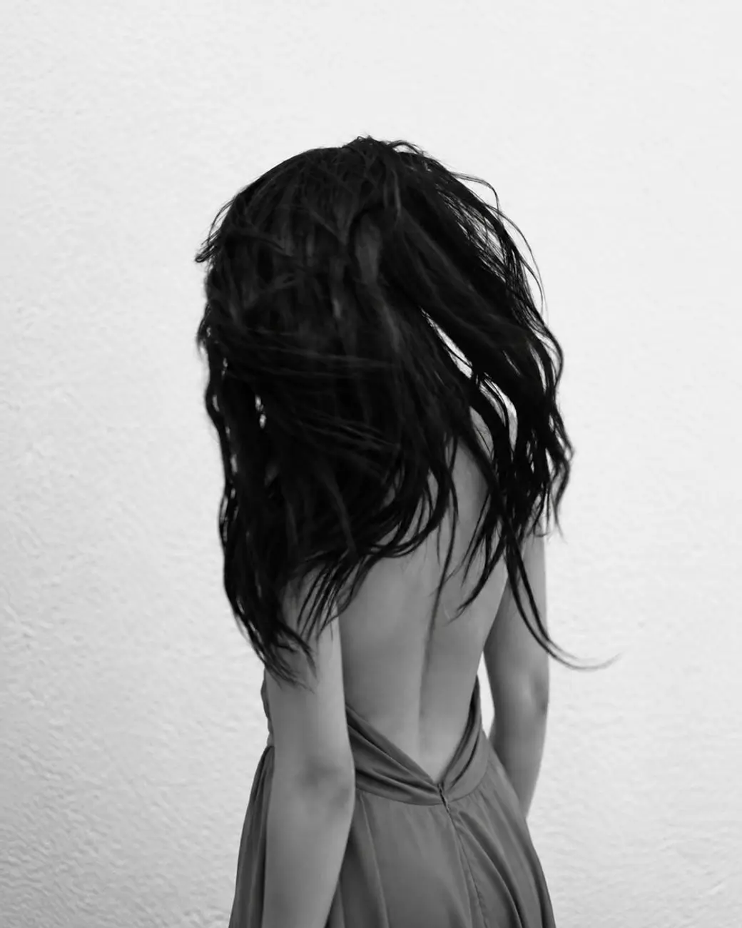 黑白照片：一名女性背對鏡頭，蓬鬆的長髮被風吹起、遮住側臉，背景是素白的牆。