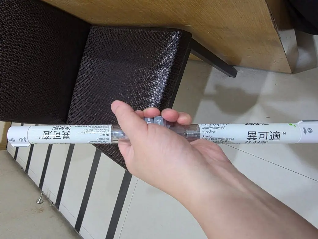 生活照片，一隻手從紙箱旁拿起一支預充填注射筆，筆身上印著中文商品名與英文學名。