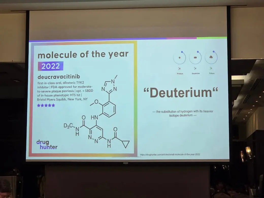 會場照片，投影幕上是一張英文投影片，左半介紹 2022 年年度分子 deucravacitinib 與其化學結構式，右半是氕、氘、氚三種同位素的示意與 Deuterium 的說明。
