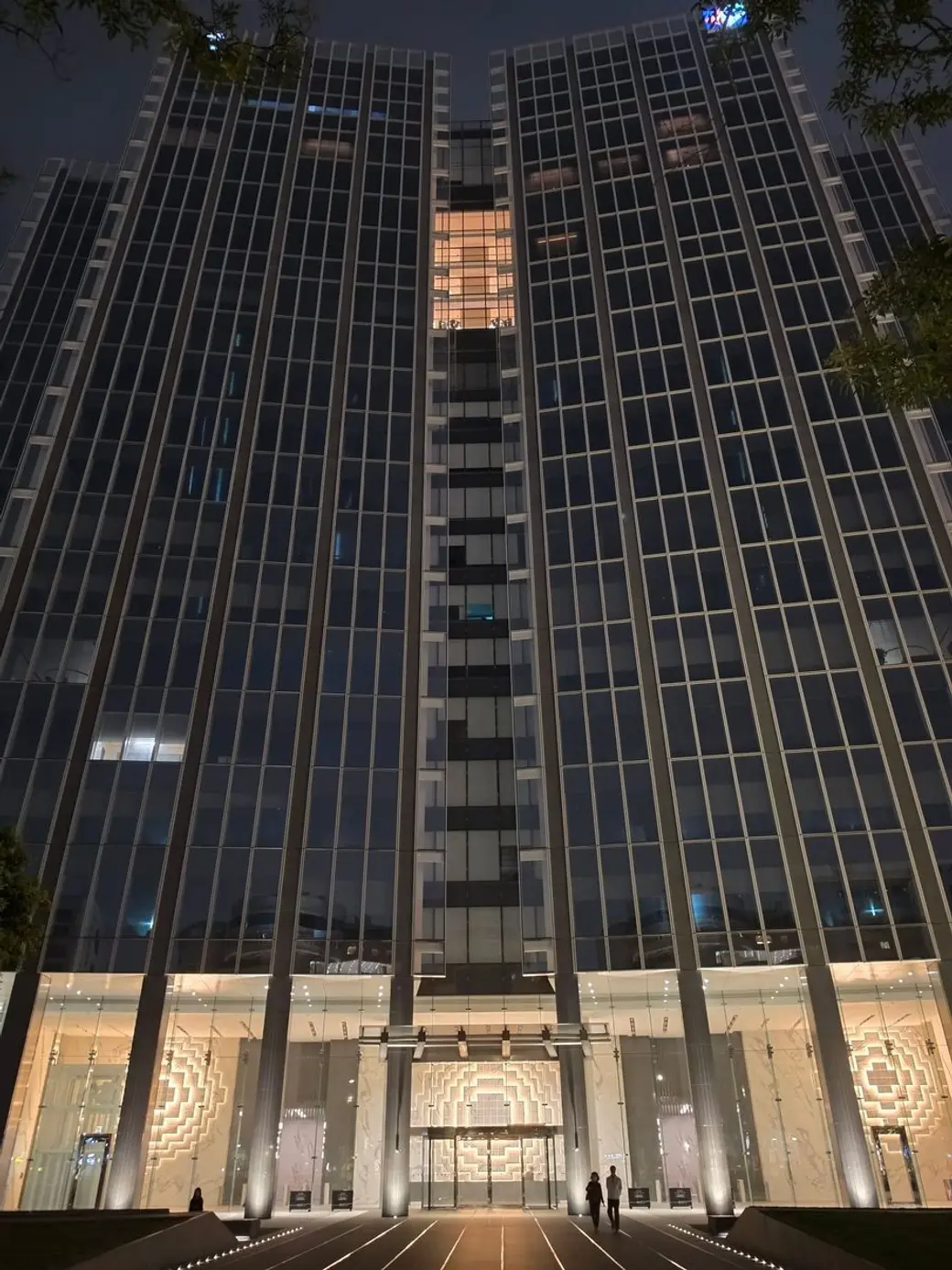 夜景照片，仰角拍攝一棟玻璃帷幕高樓的正面，入口處亮著暖色燈光，兩個人影走在前方廣場。