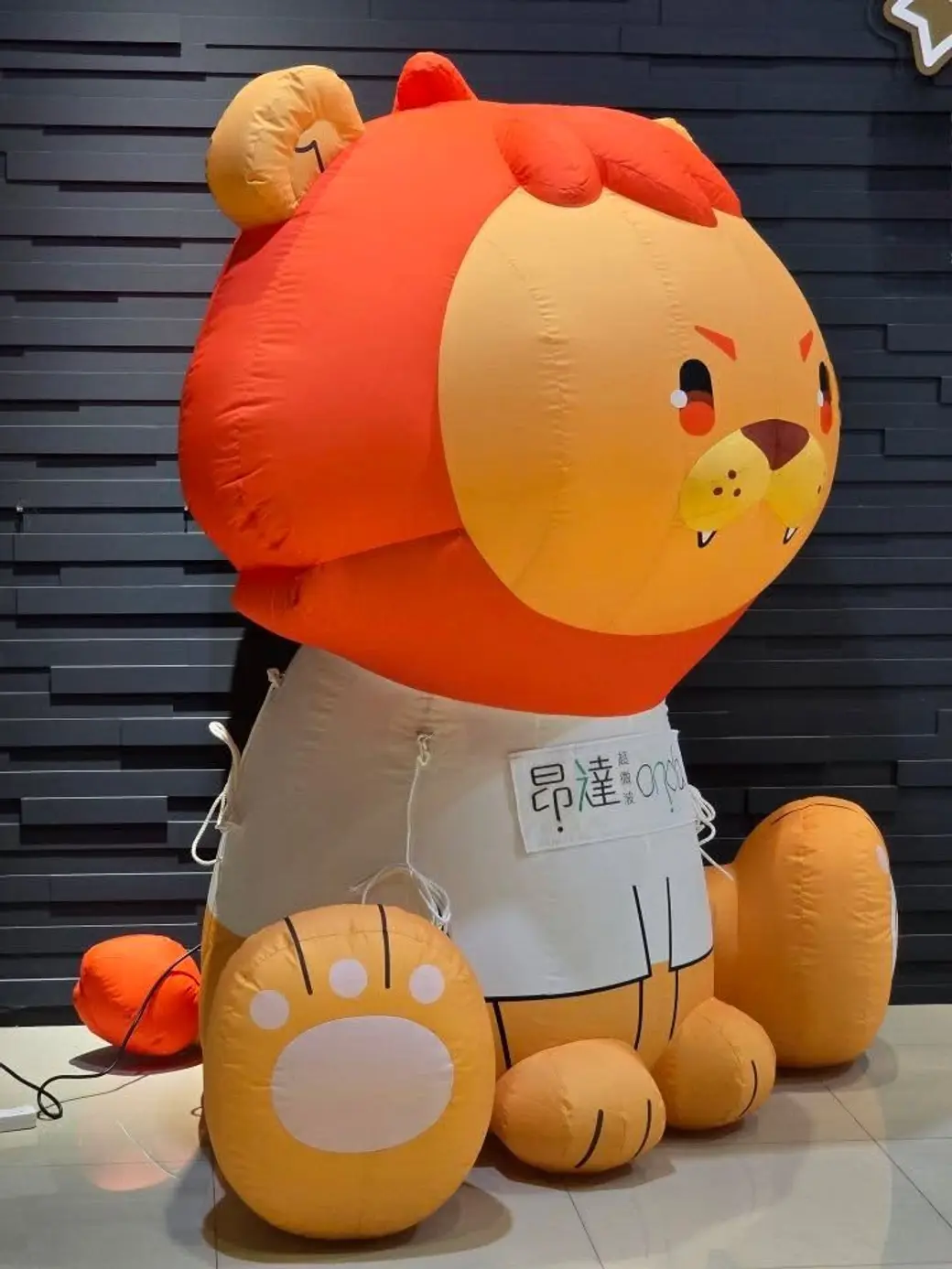 活動照片，室內展場地上立著一個巨大的充氣獅子玩偶，圍著白色圍裙，上面印著產品名稱。