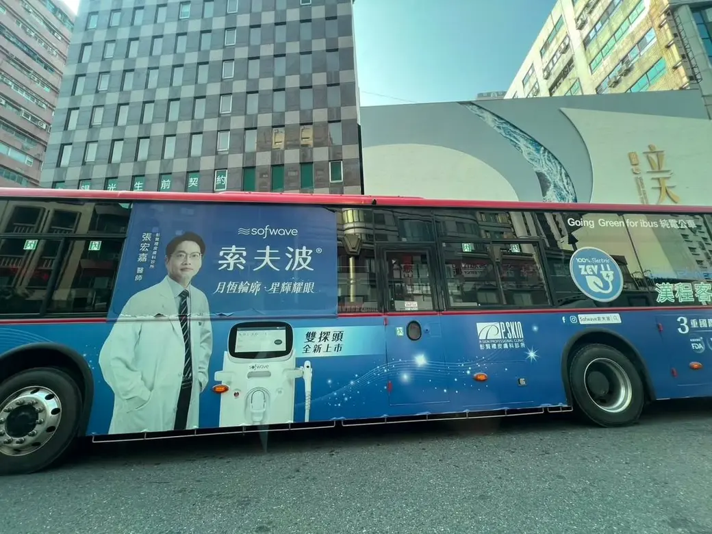 街景照片，一輛藍色公車的側身廣告，上面是一位白袍男性與一台儀器的圖像和產品名稱，背景是街邊大樓。