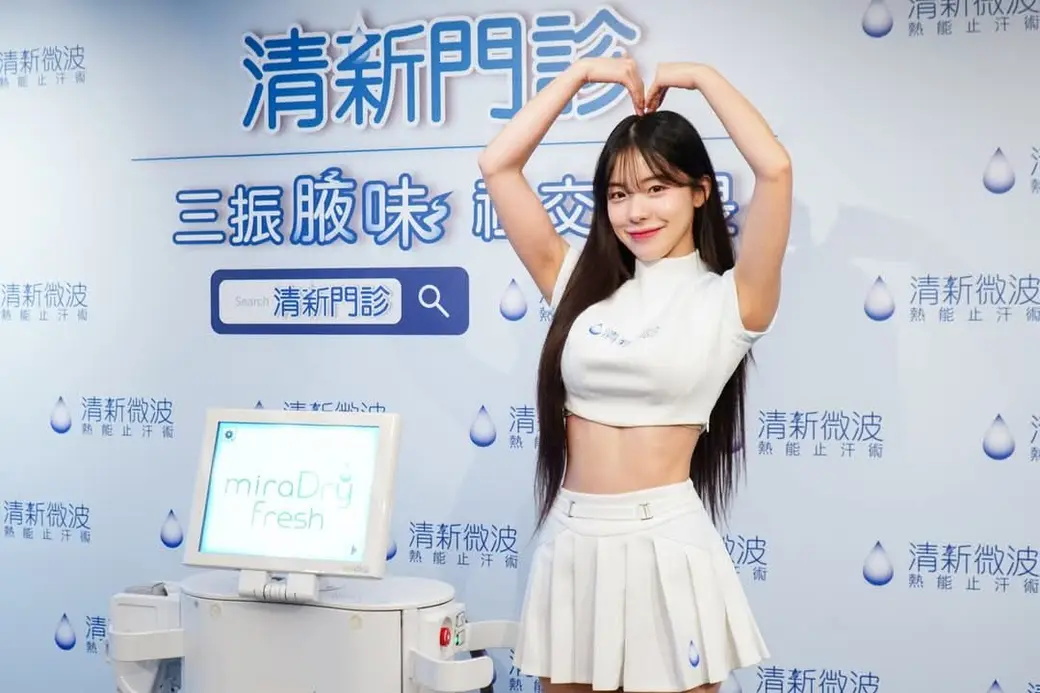 活動照片，一位女性站在藍色背板前雙手在頭頂比心，旁邊是一台微波止汗儀器，背板上重複印著產品名稱。