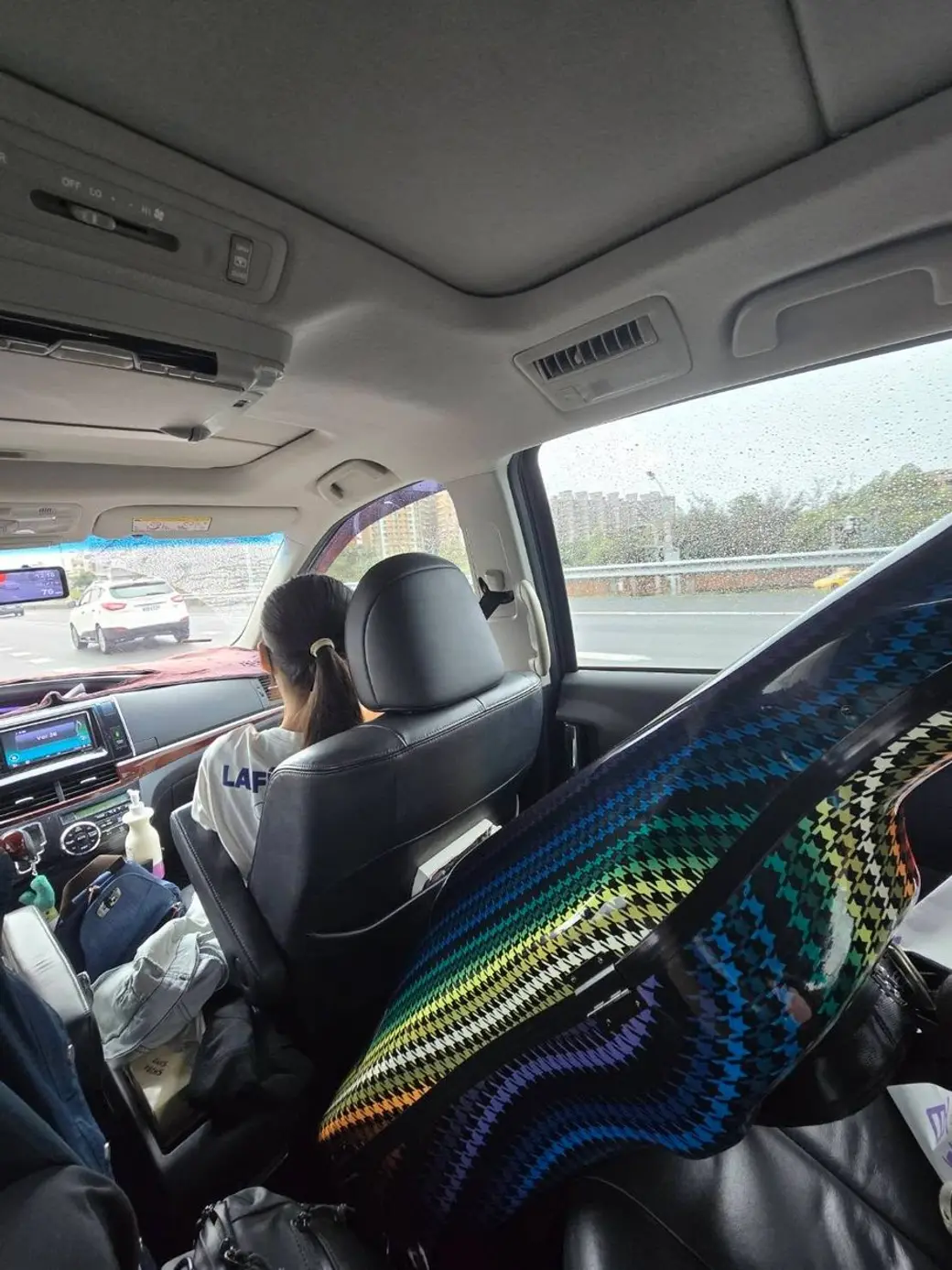車內照片，副駕駛座上有人背對鏡頭坐著，中間座椅上攤著一條彩色格紋毯子，車窗外在下雨。
