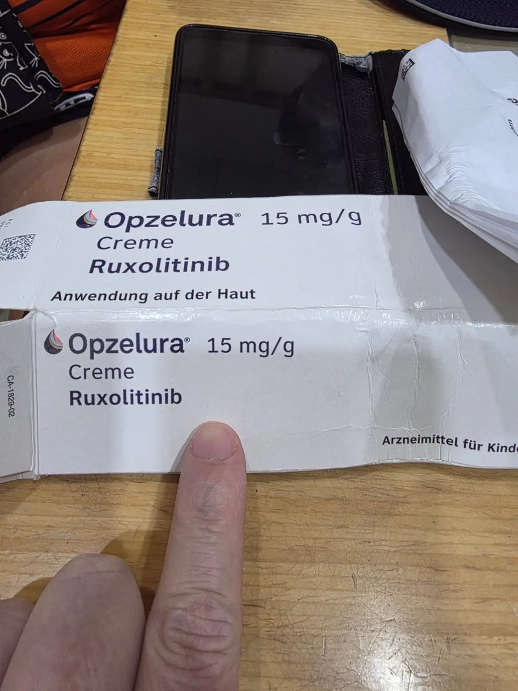 照片，桌上放著兩盒德文包裝的外用 JAK 抑制劑藥膏，一隻手的食指指著其中一盒。