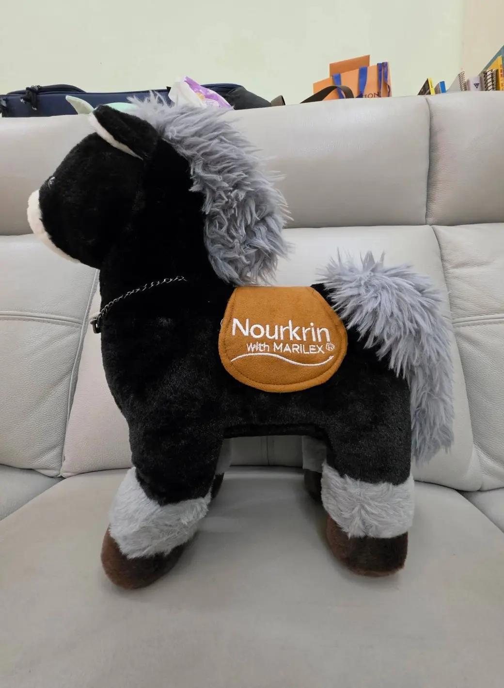 一隻黑色絨毛馬玩偶站在淺色沙發上，背上的橘色鞍墊繡著 Nourkrin with MARILEX。